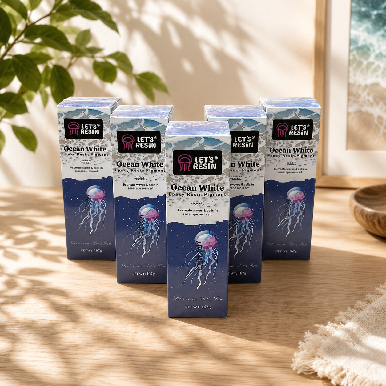 A collection of five boxes of Let's Resin Ocean White Pigment liquid ink display containers featuring jellyfish illustrations, arranged neatly on a wooden table next to a decorative plant.