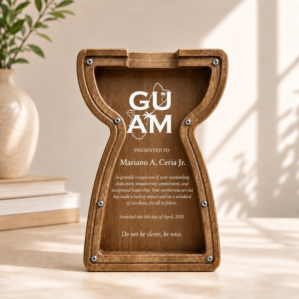 A wooden hourglass-shaped plaque with engraved text, displayed on a tabletop in a warm indoor setting with sunlight and a vase in the background.