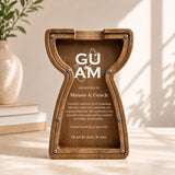 A wooden hourglass-shaped plaque with engraved text, displayed on a tabletop in a warm indoor setting with sunlight and a vase in the background.