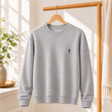 A light gray crewneck sweatshirt with a small black cross embroidered on the left chest, hanging on a wooden hanger from a wooden clothing rack in a bright, sunlit room.