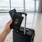 A hand holding a custom black leather passport book cover and matching luggage tag engraved with a seal logo, held up against a rolling suitcase handle in an airport terminal.