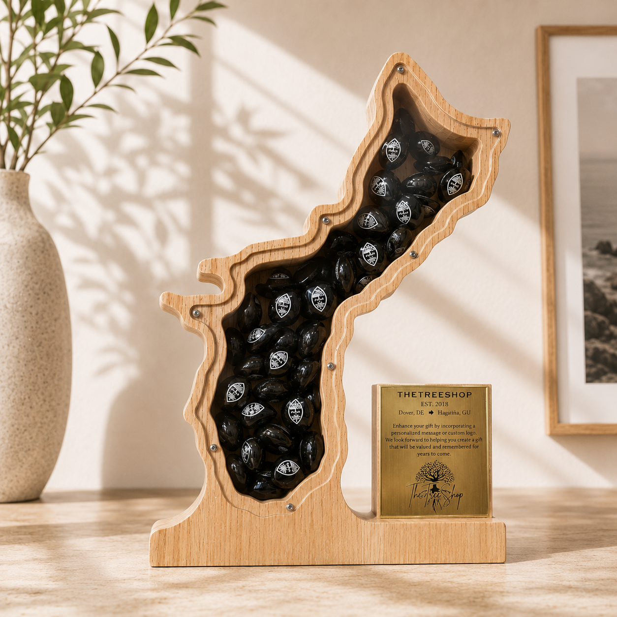 A large 12-inch custom wooden shadow box plaque carved into the geographical silhouette of Guam Island, featuring a clear acrylic front window filled with small black decorative stones, paired alongside a personalized engraved gold metal trophy plate on a light wood studio table.
