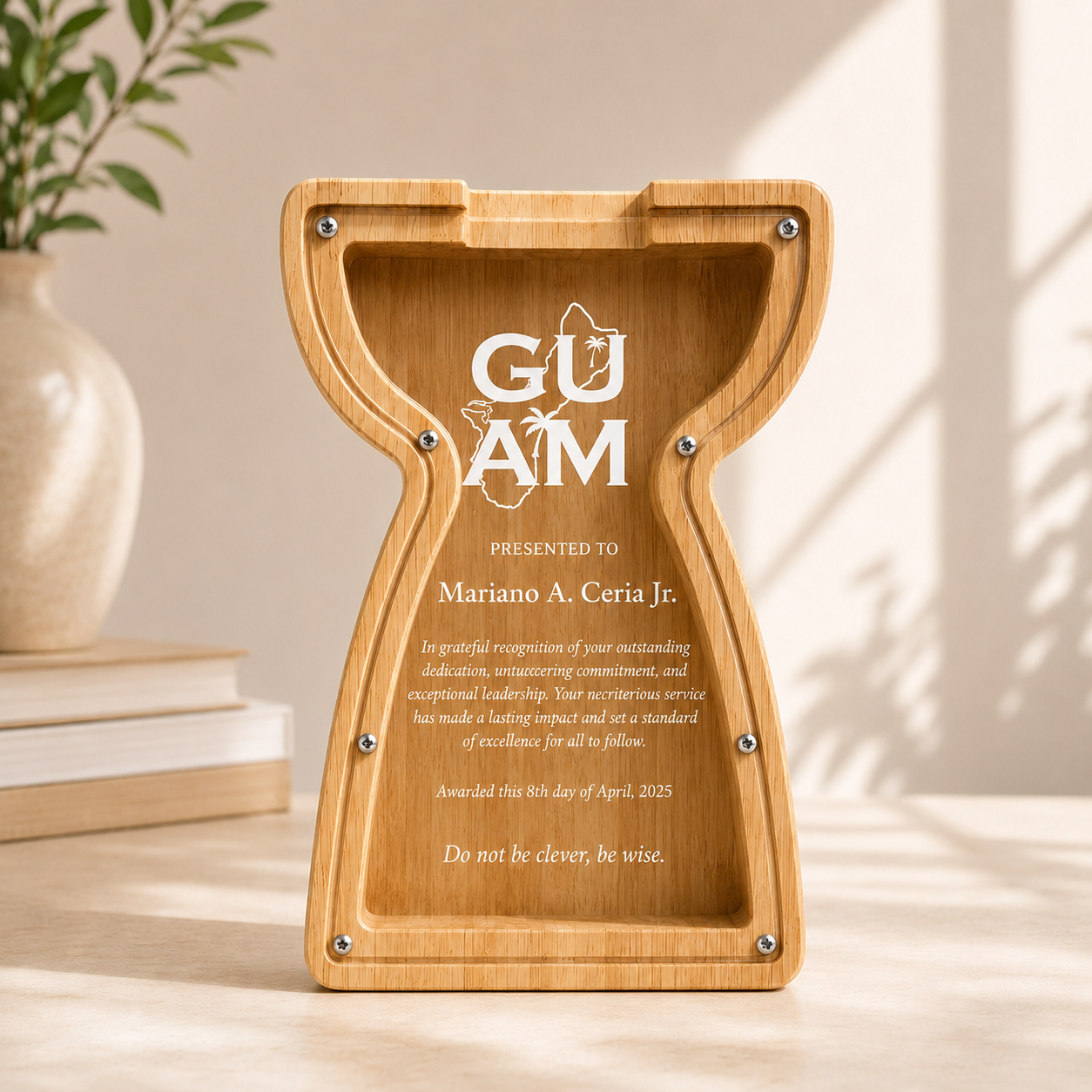 A compact 7.5-inch custom laser-engraved wooden piggy bank and commemorative award carved in a Guam latte stone shape with a clear acrylic window front panel, displayed on a wooden desk next to a stack of books and a white vase.