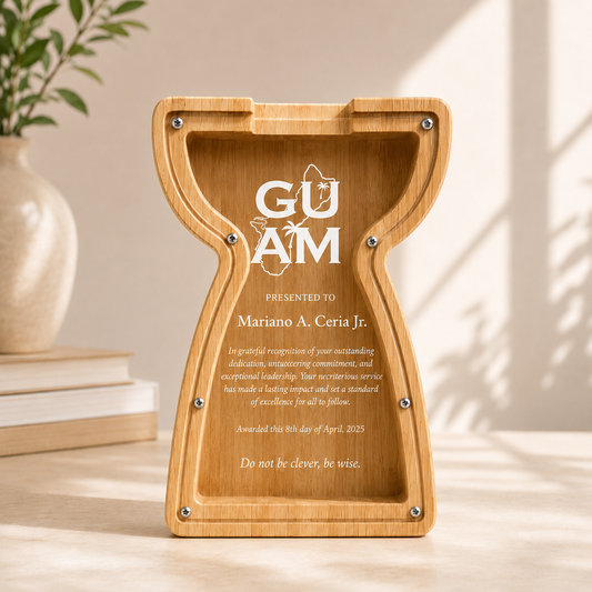 A compact 7.5-inch custom laser-engraved wooden piggy bank and commemorative award carved in a Guam latte stone shape with a clear acrylic window front panel, displayed on a wooden desk next to a stack of books and a white vase.