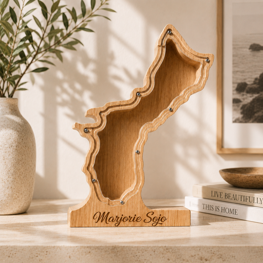 A custom laser-engraved wooden piggy bank carved into the geographical silhouette of Guam Island, featuring a clear acrylic front window and a solid base personalized with the name 'Marjorie Sojo' on a bright studio table.