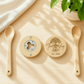 A top-down view of two round custom wood bottle openers displaying the metal insert and a personalized 'The Tree Shop' engraving, styled on a bright table background next to a white fabric cloth and wooden cooking spoons.
