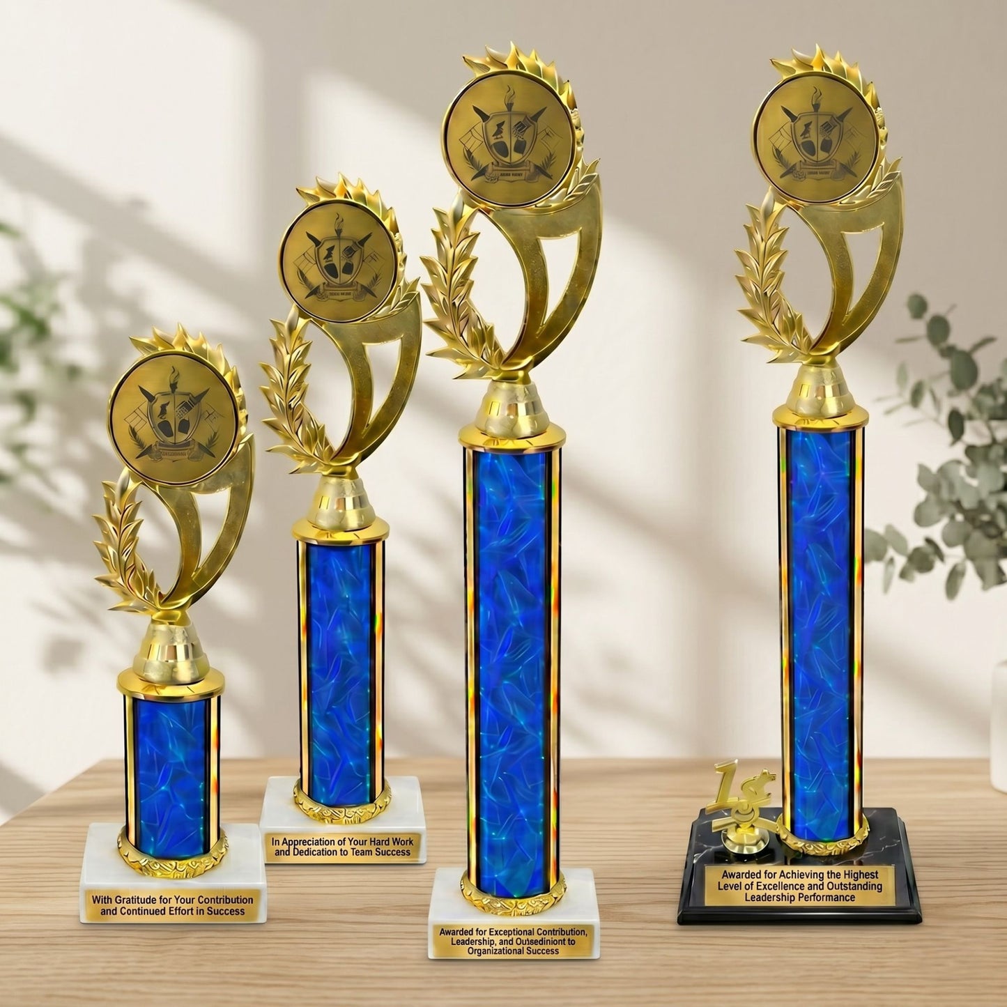 A lineup of four customizable classic single column trophies in varying heights featuring textured blue columns, shiny gold trim accents, white and black marble bases with personalized engraved nameplates, and gold circular trim toppers on a wooden table.