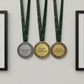 A side-by-side wall display of three custom personalized achievement medals in silver, gold, and bronze hanging by their dark green neck ribbons against a clean white backdrop.