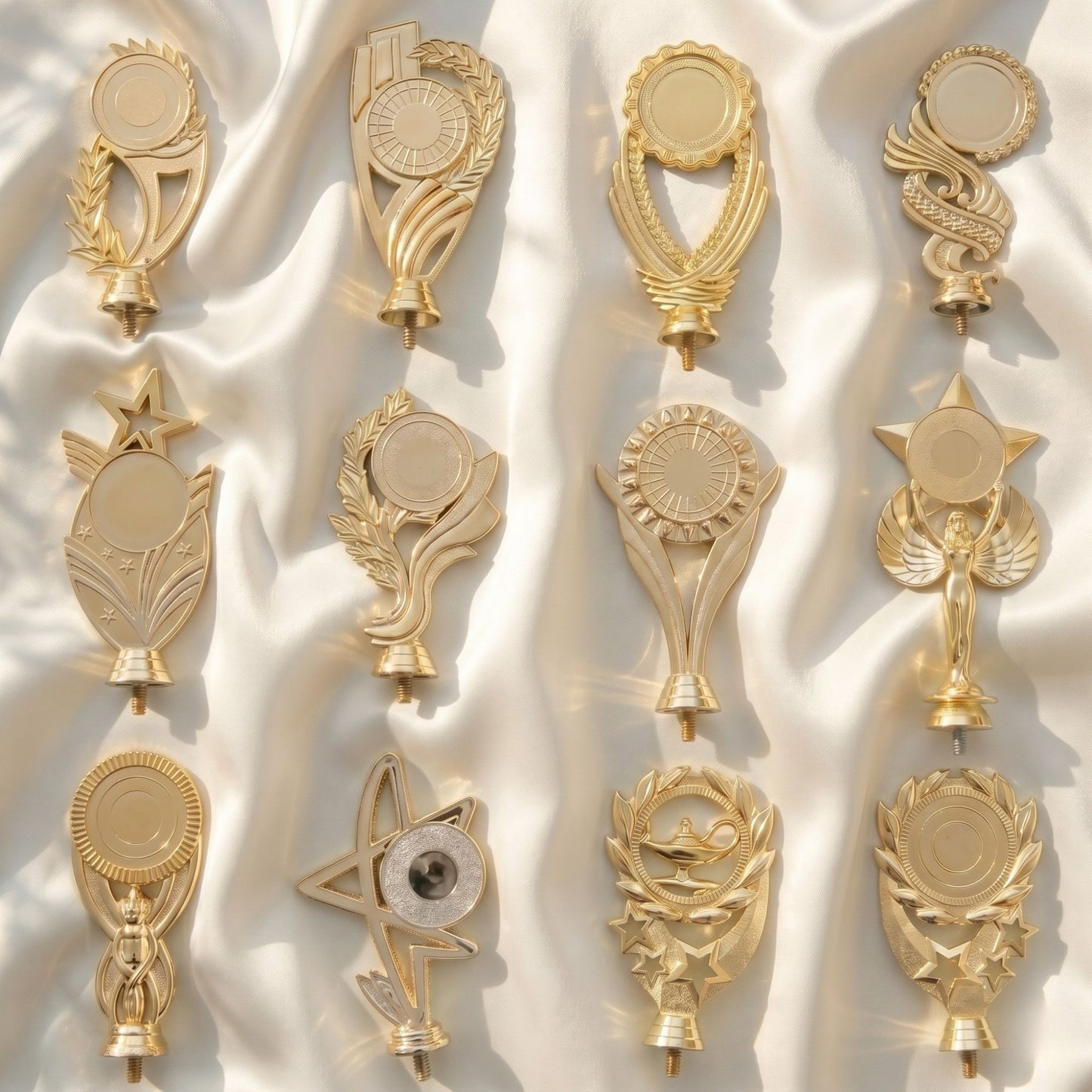 A collection of twelve different personalized brilliant gold trophy toppers featuring stars, victory wreaths, and classic academic motifs arranged on a soft white satin fabric background.