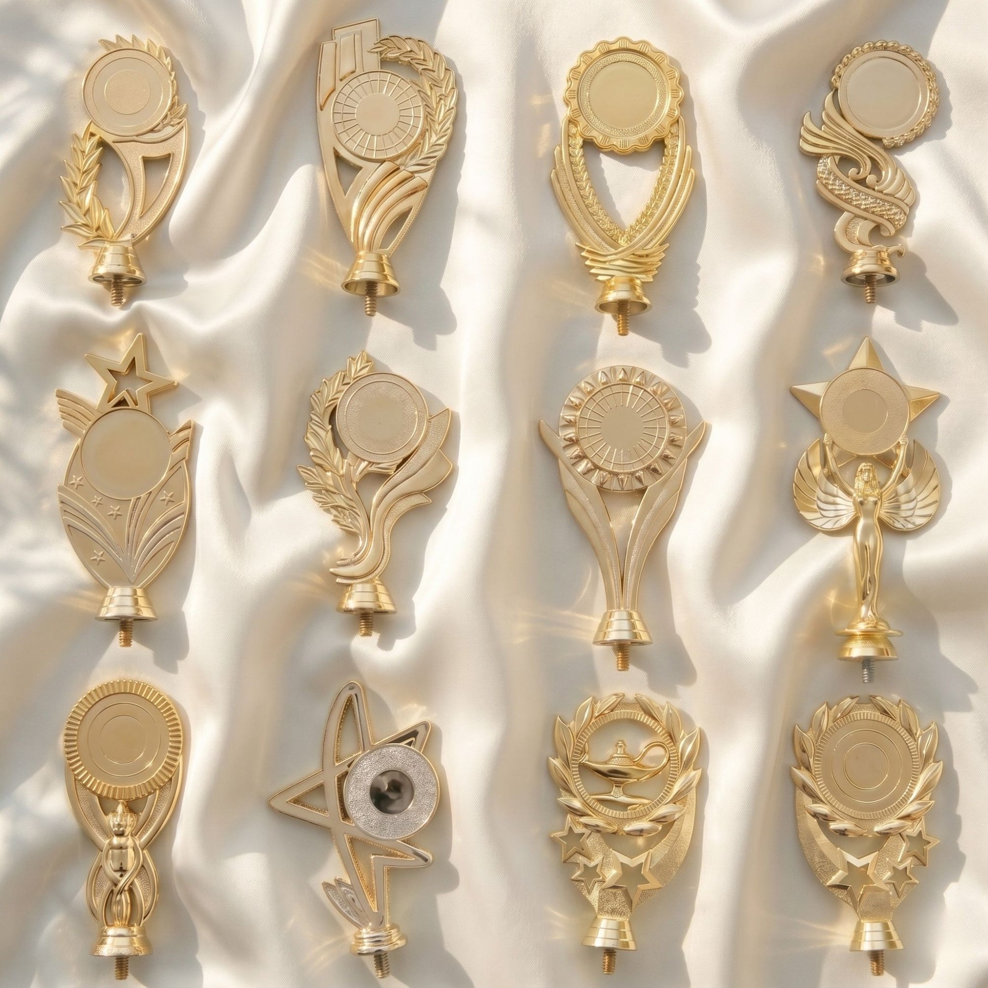 A collection of twelve different personalized brilliant gold trophy toppers featuring stars, victory wreaths, and classic academic motifs arranged on a soft white satin fabric background.