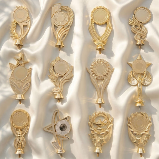 A collection of twelve different personalized brilliant gold trophy toppers featuring stars, victory wreaths, and classic academic motifs arranged on a soft white satin fabric background.