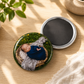Close-up of a 2.25 inch custom round button magnet featuring a glossy sleeping baby portrait photo, shown next to its black magnetic back side on a wooden tabletop with soft leaf shadows.