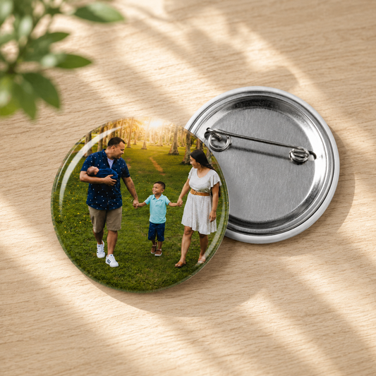 Close-up view of a 2.25 inch custom round button pin displaying a personalized glossy family outdoor portrait photo, shown alongside the blank silver metal pinback side on a light wood table with natural leaf shadows.
