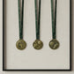 A minimalist shadow box wall display showcasing three personalized antique bronze sports medals hanging neatly by their green neck ribbons against a soft beige textured backdrop.