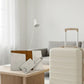 An elegant white hard-case rolling suitcase standing in a bright minimalist hotel room corner, with a matching customized leather passport wallet pack resting nearby on an accent bench.