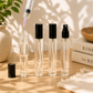 A set of four clear 10ml glass fine mist spray bottles with black atomizers and caps standing on a wooden table, with a syringe filling one bottle near a decorative beige vase.