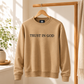 Close-up of a premium sand tan colored crewneck pullover sweater showing clean white direct embroidery text that reads 'TRUST IN GOD'.