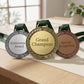 Three custom-engraved zinc alloy award medallions in antique silver, bright gold, and classic bronze with laurel leaf borders and green patterned neck ribbons, standing on a light wood display desk.