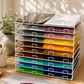 A white wire tabletop storage rack filled with colorful 15x12 inch Siser EasyWeed heat transfer vinyl sheets in red, yellow, orange, blue, and green on a wooden craft desk next to a potted plant.