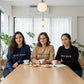 Three women sitting together at a wooden table inside a bright room, smiling and wearing the custom embroidered 'TRUST IN GOD' cotton crewneck sweaters in navy blue, sand tan, and classic black.