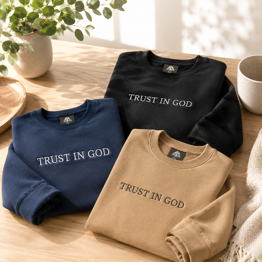 A collection of three premium cotton crewneck sweaters with 'TRUST IN GOD' custom embroidery across the chest, displayed in navy blue, classic black, and sand tan folded neatly on a wooden display table.