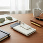 A personalized white leather passport holder layed flat on a warm wooden work desk next to a notebook, sunglasses, and a hot white ceramic mug of coffee.